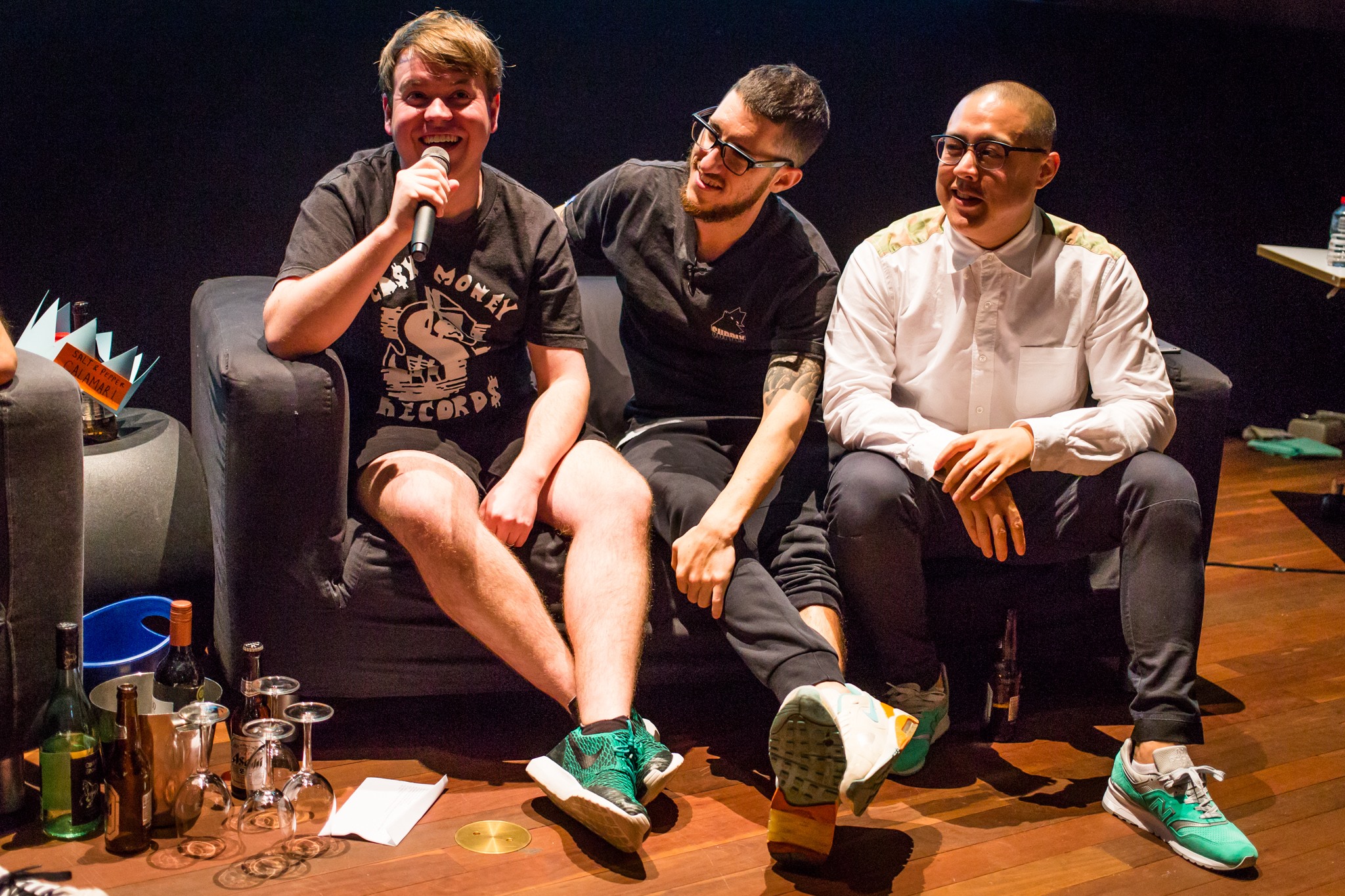 Three men on a couch, one is holding a microphone presenting a live podcast