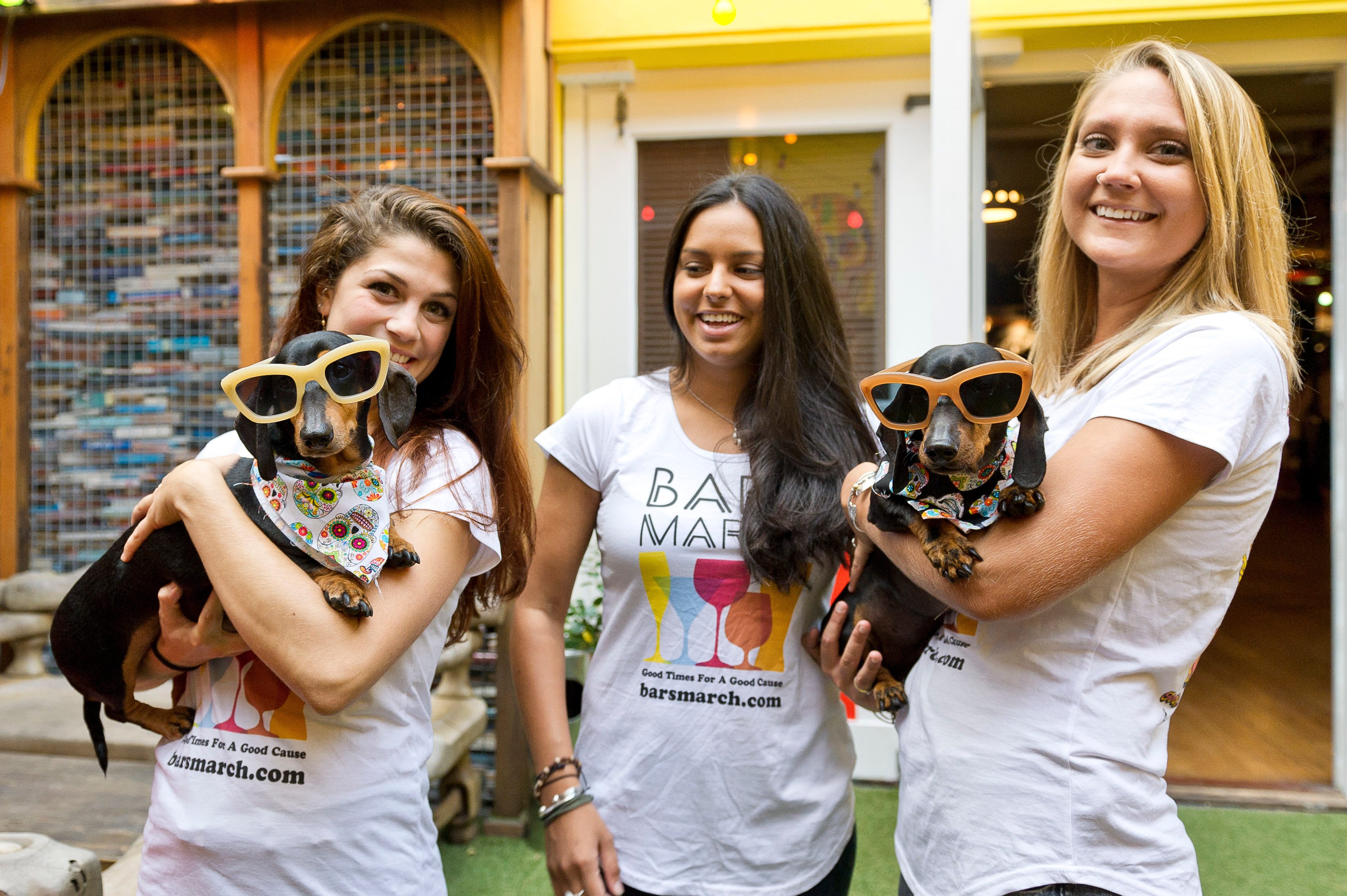 Three women, two are holding sausage dogs with oversizes sunglasses