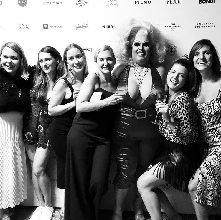 A group of people including drag queen Maxi Shield posing in front of a wall