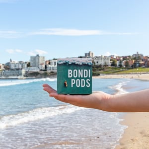 A hand holds a box of Bondi Pods coffee capsules in front of the Bondi Beach coastine