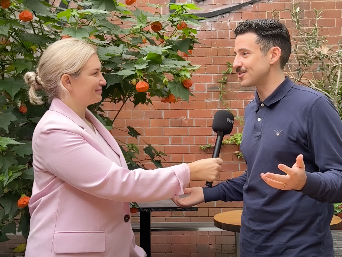 Naomi Ross, agency director at Distil, holds a microphone as she interviews Andrew Knox, hospitality market research expert. There is a brick wall and a plant with flowers behind them.