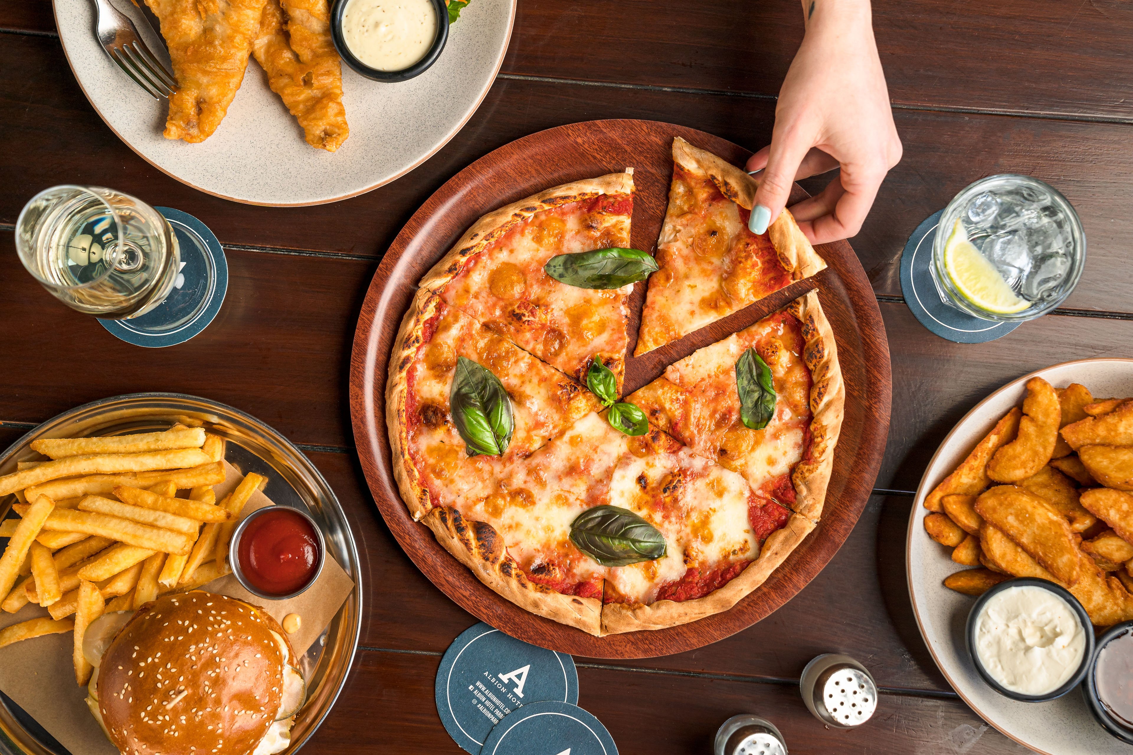 Top view of a hand taking a slice of pizza, surrounded by other food and drink