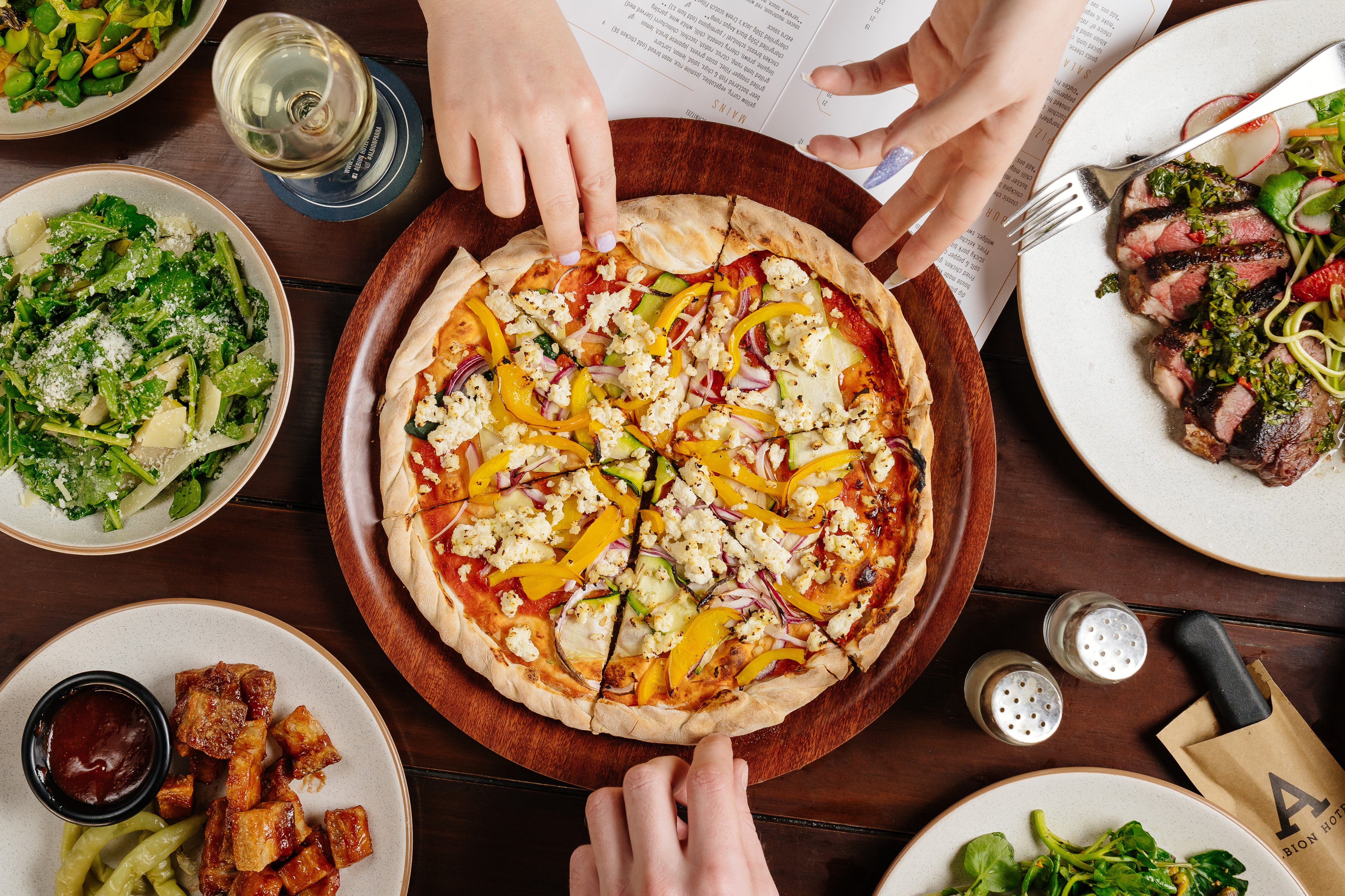 Flatlay of pizza surrounded by side dishes, with hands reaching for a slice